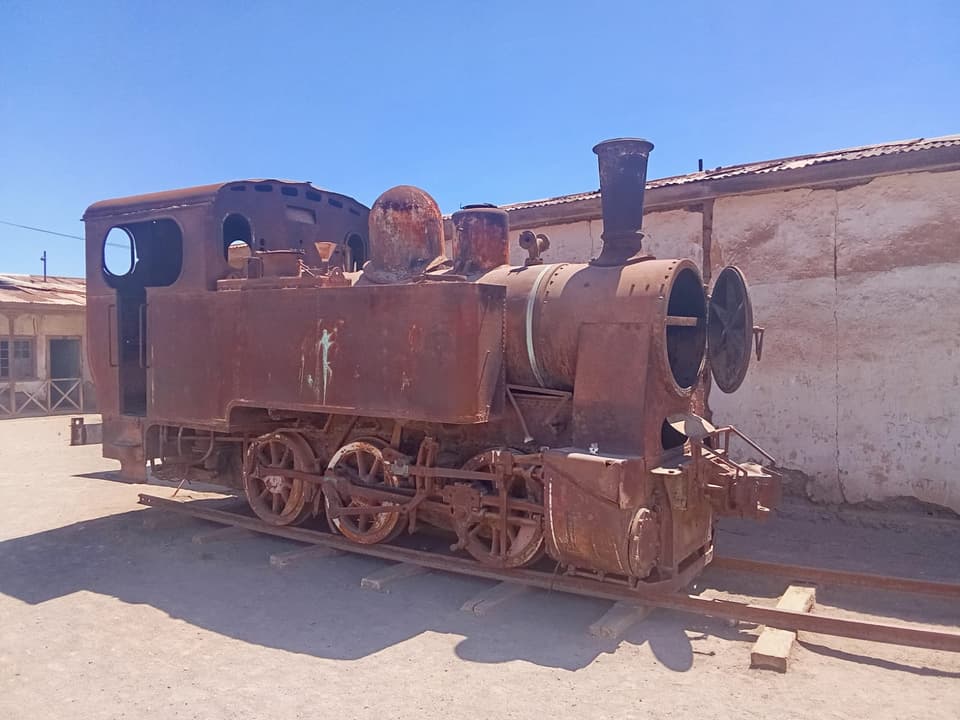 Locomotora a vapor oxidada sobre rieles, frente a un muro de adobe